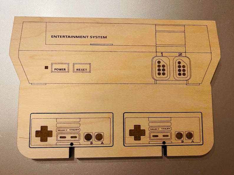 K&ouml;nnte beinhalten: Ein Holzausschnitt einer klassischen Videospielkonsole mit der Aufschrift "ENTERTAINMENT SYSTEM". Das Design umfasst Controller-Anschl&uuml;sse, Power- und Reset-Tasten sowie zwei Controller. Das Holz hat eine helle, nat&uuml;rliche Oberfl&auml;che.