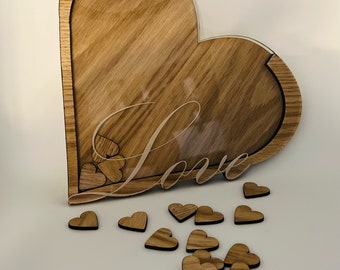 Könnte beinhalten: Eine herzförmige Holzkiste mit einer durchsichtigen Acrylglasscheibe vorne. Die Kiste ist mit dem Wort "Love" auf der Vorderseite graviert. Kleine Holzh Herzen sind um die Kiste herum verstreut.