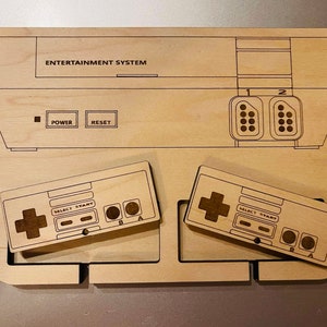 K&ouml;nnte beinhalten: Ein Holz-Organizer im NES-Design mit ge&auml;tzten Details. Das Design umfasst den Umriss der Konsole mit der Aufschrift "ENTERTAINMENT SYSTEM" und zwei Controllern. Die Controller haben Richtungspads, Select-, Start- und A/B-Tasten.