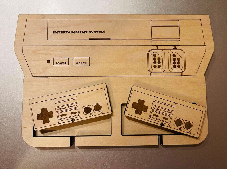 K&ouml;nnte beinhalten: Ein Holz-Organizer im NES-Design mit ge&auml;tzten Details. Das Design umfasst den Umriss der Konsole mit der Aufschrift "ENTERTAINMENT SYSTEM" und zwei Controllern. Die Controller haben Richtungspads, Select-, Start- und A/B-Tasten.