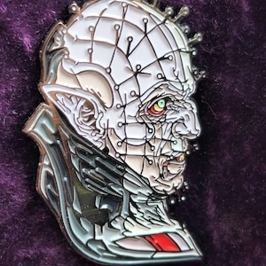 May include: A silver and white enamel pin featuring a detailed illustration of a horror movie character with a stitched-up face and a sharp, pointed nose. The character has a menacing expression and is wearing a dark, hooded cloak.