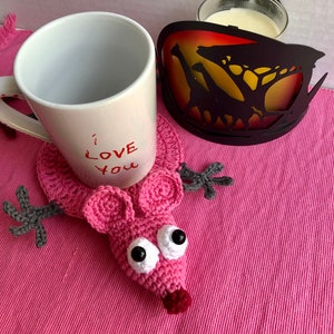 May include: A white mug with "I Love You" in red lettering sits on a pink crocheted mouse-shaped coaster. A decorative coaster with a giraffe silhouette and a glass of liquid are also present. The items rest on a pink table runner.