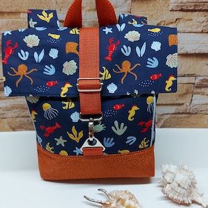 May include: A small blue backpack with an orange strap and a sea life print. The print features seahorses, jellyfish, starfish, and an octopus. The backpack has a flap that folds over the top and a buckle closure.