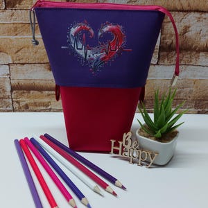 Op de afbeelding: Tweekleurige etui met een hartvormig ontwerp en een ritssluiting. De etui is paars en rood, met een hartafbeelding met muzieknoten en een hartslaglijn. Er zijn verschillende kleurpotloden voor de etui geplaatst.