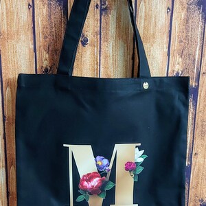 Op de afbeelding: Zwarte canvas tote bag met een gouden letter "M" versierd met kleurrijke bloemen.