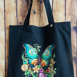 Könnte beinhalten: Schwarze Canvas-Tragetasche mit einem bunten Schmetterlings- und Blumendesign. Der Schmetterling hat grüne, gelbe und orange Flügel. Die Blumen sind rosa, gelb und weiß.