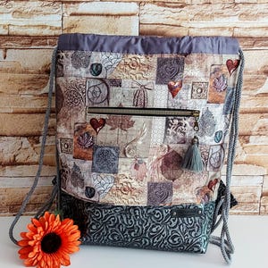 Peut inclure: Un sac à dos à cordon de serrage en patchwork avec un dessus et des bretelles gris. Le corps principal présente un collage de carrés avec des motifs de cœur, de feuilles et de fleurs dans des tons de marron, beige et gris. Une poche zippée et un pompon gris ajoutent des détails.
