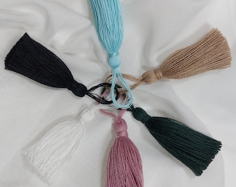 May include: A collection of six decorative tassels arranged in a circular pattern. The tassels are colored light blue, tan, dark green, dusty rose, white, and black. Each tassel features a tied top loop and a dense fringe of fine thread strands.