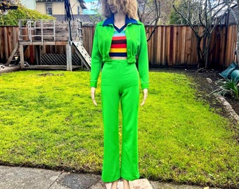 May include: A vibrant green two-piece outfit. The jacket has a blue collar and a multi-colored striped top underneath. The pants are high-waisted and flared. The outfit is displayed on a mannequin in a grassy outdoor setting.