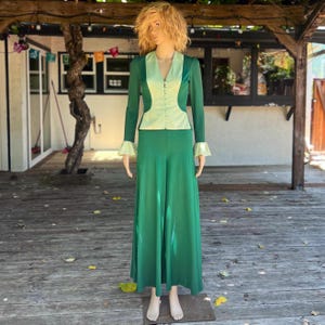 May include: A green two-piece suit on a mannequin. The suit features a long-sleeved jacket with a light green panel and matching wide-leg pants. The jacket has a V-neck and button closure. The cuffs of the jacket and the lapel are light green.