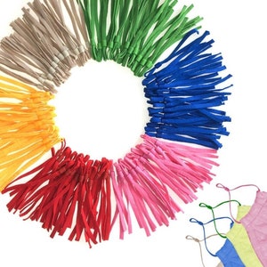 May include: Assortment of colorful elastic straps for face masks. The straps are arranged in a circle with the colors beige, green, blue, pink, red, and yellow. There are three face masks in the bottom right corner of the image, one blue, one yellow, and one pink.