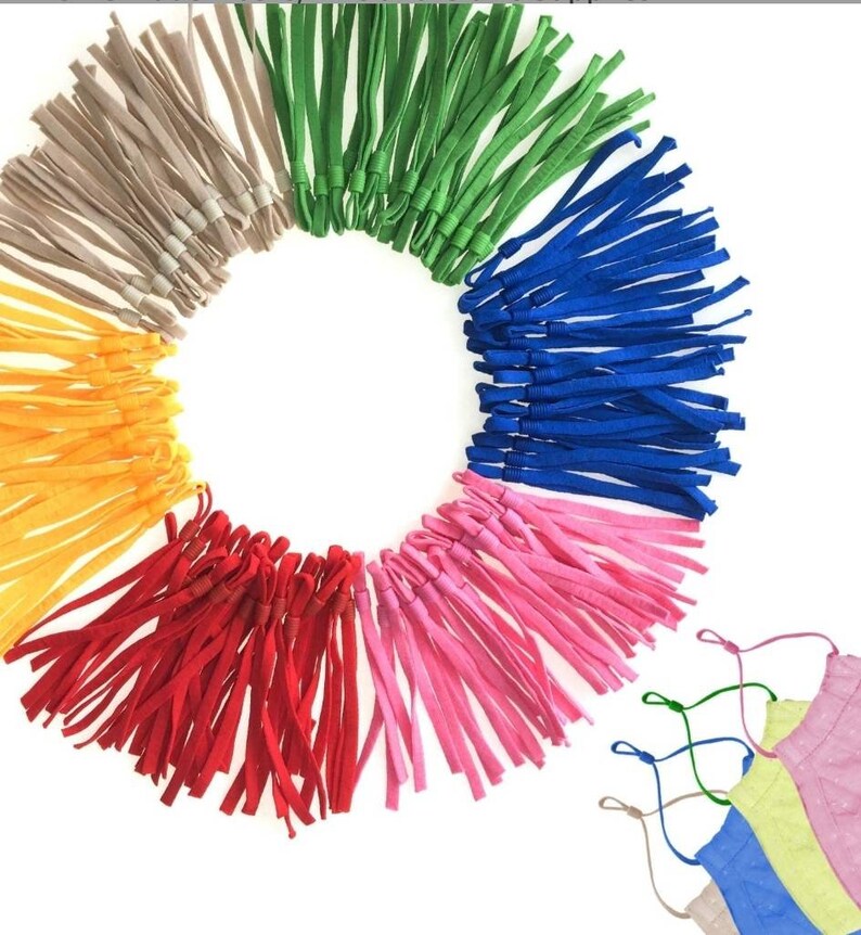 May include: Assortment of colorful elastic straps for face masks. The straps are arranged in a circle with the colors beige, green, blue, pink, red, and yellow. There are three face masks in the bottom right corner of the image, one blue, one yellow, and one pink.