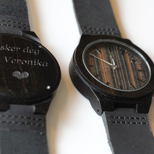 Può includere: Due orologi da polso in legno nero con cinturini in pelle nera. Un orologio ha un quadrante in legno con un motivo a venature di legno nero e marrone. L'altro orologio ha un quadrante in legno nero con l'iscrizione "Elsker deg din Veronika" e un piccolo cuore.