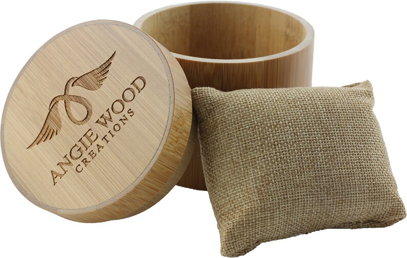 May include: A round wooden box with a lid. The lid has a logo that says "Angie Wood Creations" with a stylized bird design. The box is sitting on a white background next to a small burlap pouch.