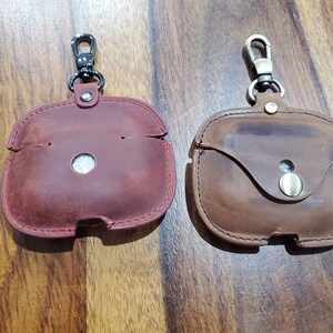 May include: Two leather cases with keychains for wireless earbuds. The case on the left is red and the case on the right is brown.