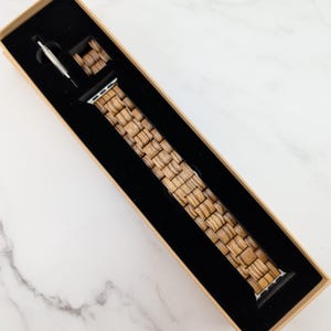 May include: A wooden watch band in a brown cardboard box with a black interior. The band is made of woven wood and has a silver buckle.