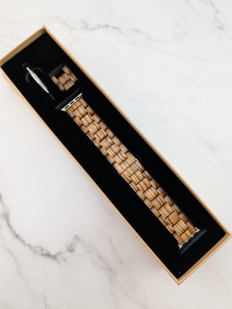 May include: A wooden watch band in a brown cardboard box with a black interior. The band is made of woven wood and has a silver buckle.