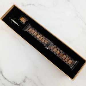 May include: A brown wooden watch band in a box. The band is made of multiple pieces of wood that are connected together. The band is designed to fit a smartwatch.