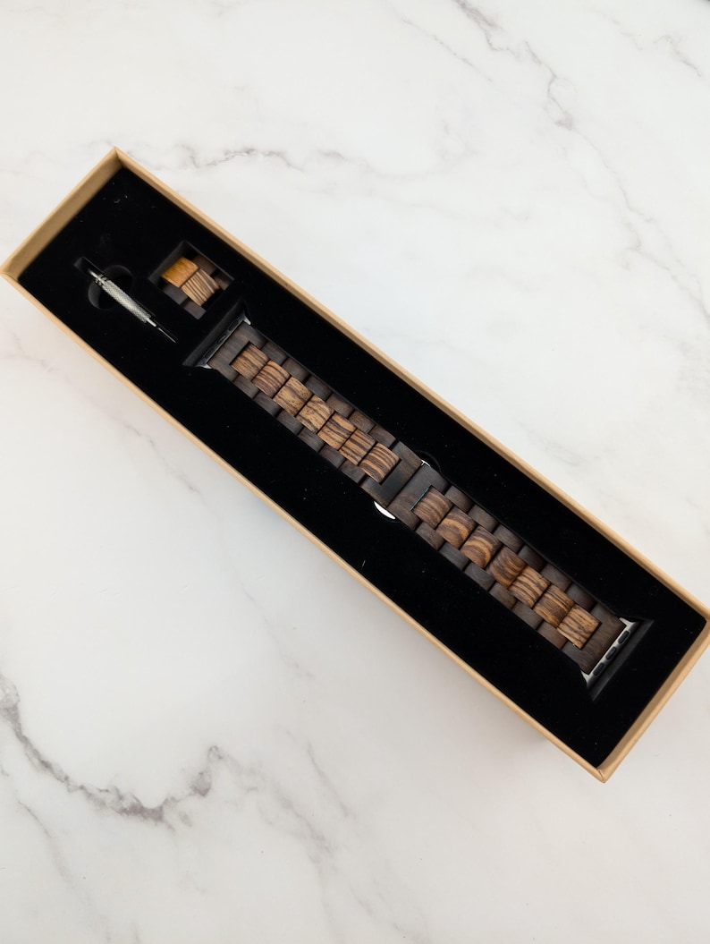 May include: A brown wooden watch band in a box. The band is made of multiple pieces of wood that are connected together. The band is designed to fit a smartwatch.