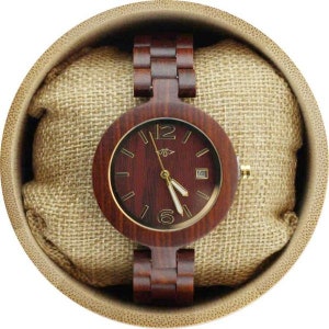 May include: A wooden wristwatch with a brown wood band and a round, dark red wood face with gold accents. The watch is in a brown, circular box with a beige fabric lining.