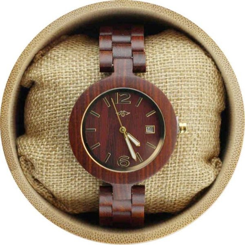 May include: A wooden wristwatch with a brown wood band and a round, dark red wood face with gold accents. The watch is in a brown, circular box with a beige fabric lining.
