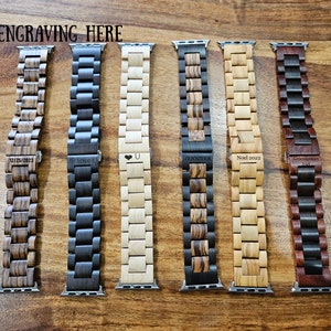 May include: Six different wooden Apple Watch bands with various engravings. The bands are made of different types of wood, including dark wood, light wood, and a combination of both. The engravings include dates, names, and a heart symbol.