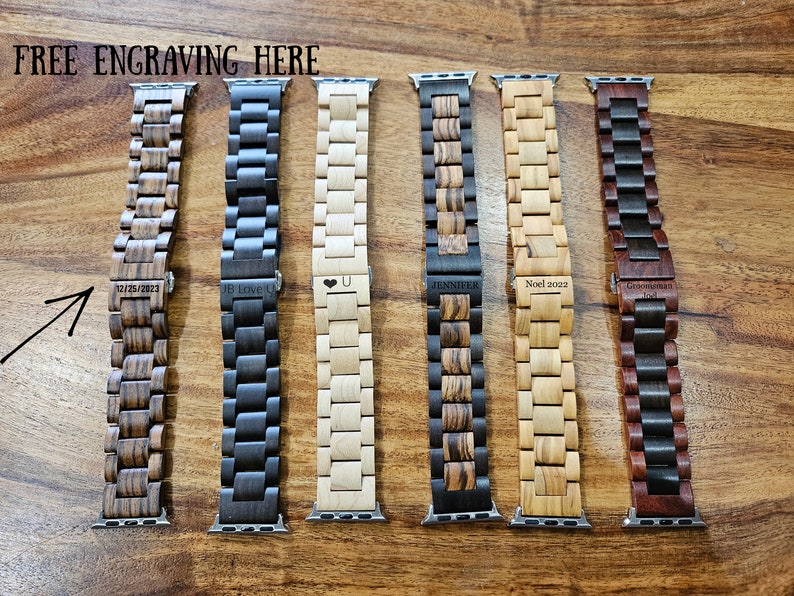May include: Six different wooden Apple Watch bands with various engravings. The bands are made of different types of wood, including dark wood, light wood, and a combination of both. The engravings include dates, names, and a heart symbol.