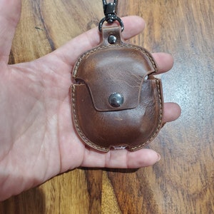 May include: A brown leather case with a metal carabiner clip for carrying wireless earbuds. The case has a small pocket on the front.