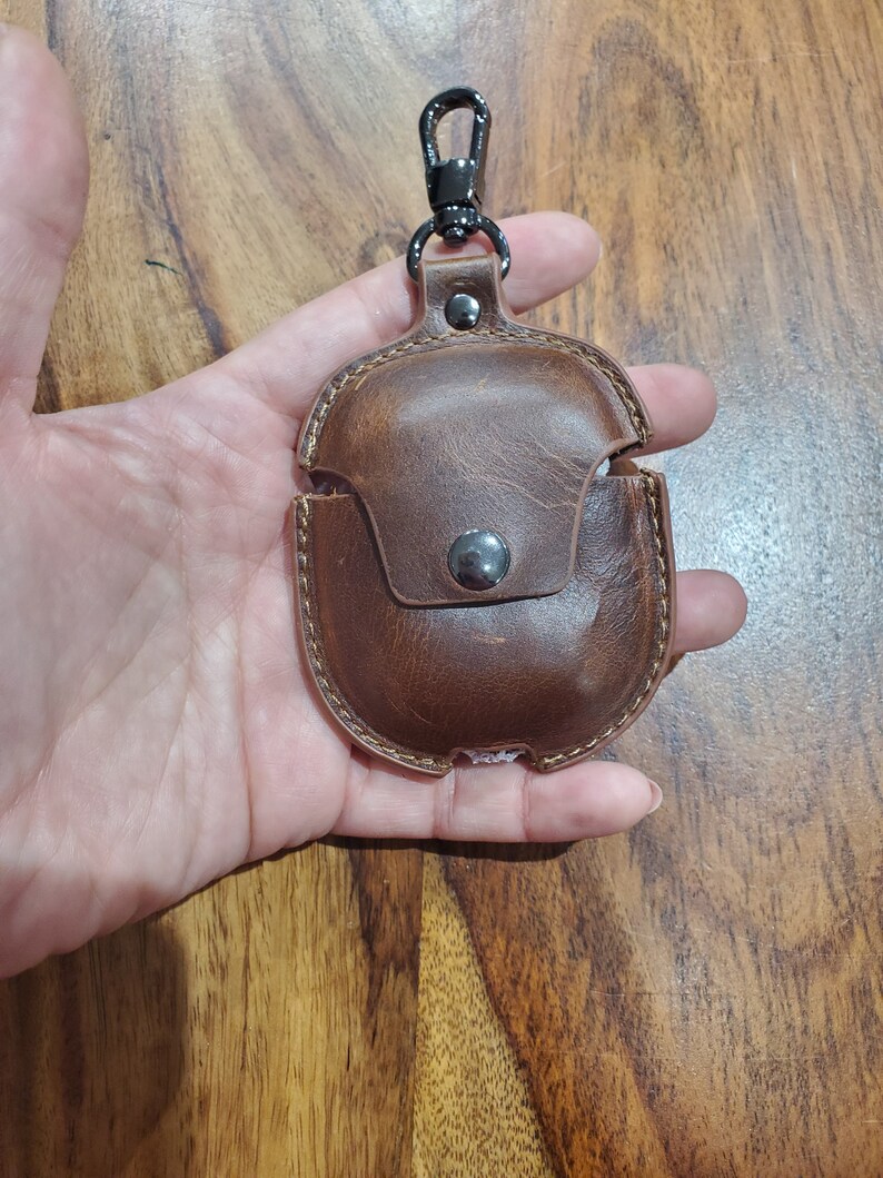 May include: A brown leather case with a metal carabiner clip for carrying wireless earbuds. The case has a small pocket on the front.