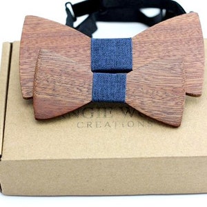 Può includere: Un papillon in legno con un nastro in denim blu. Il papillon è in una scatola marrone con il testo "Anglewood Creations".