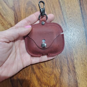 May include: A burgundy leather case with a metal clasp and a silver button. The case is designed to hold a pair of wireless earbuds.