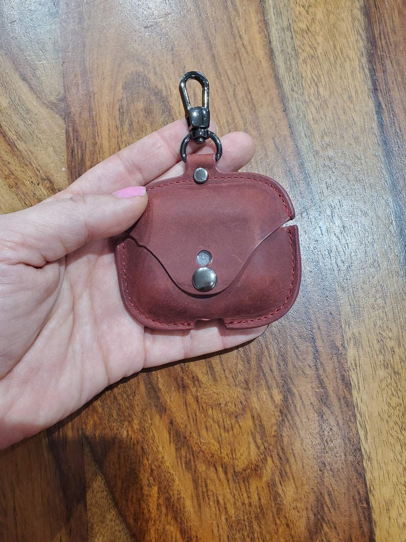May include: A burgundy leather case with a metal clasp and a silver button. The case is designed to hold a pair of wireless earbuds.