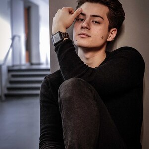 May include: A young person wearing a black long-sleeved shirt and dark blue jeans. They are sitting with their right arm resting on their left leg and their right hand touching their forehead. They are wearing a square-shaped watch with a brown band on their left wrist.