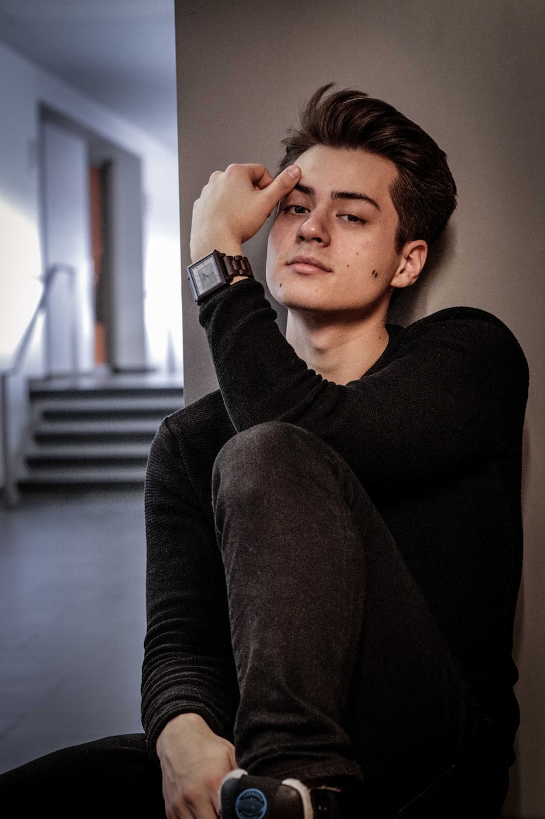 May include: A young person wearing a black long-sleeved shirt and dark blue jeans. They are sitting with their right arm resting on their left leg and their right hand touching their forehead. They are wearing a square-shaped watch with a brown band on their left wrist.