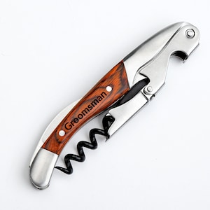 May include: A wooden and silver corkscrew with a black corkscrew and a bottle opener. The handle is engraved with the word "Groomsman".