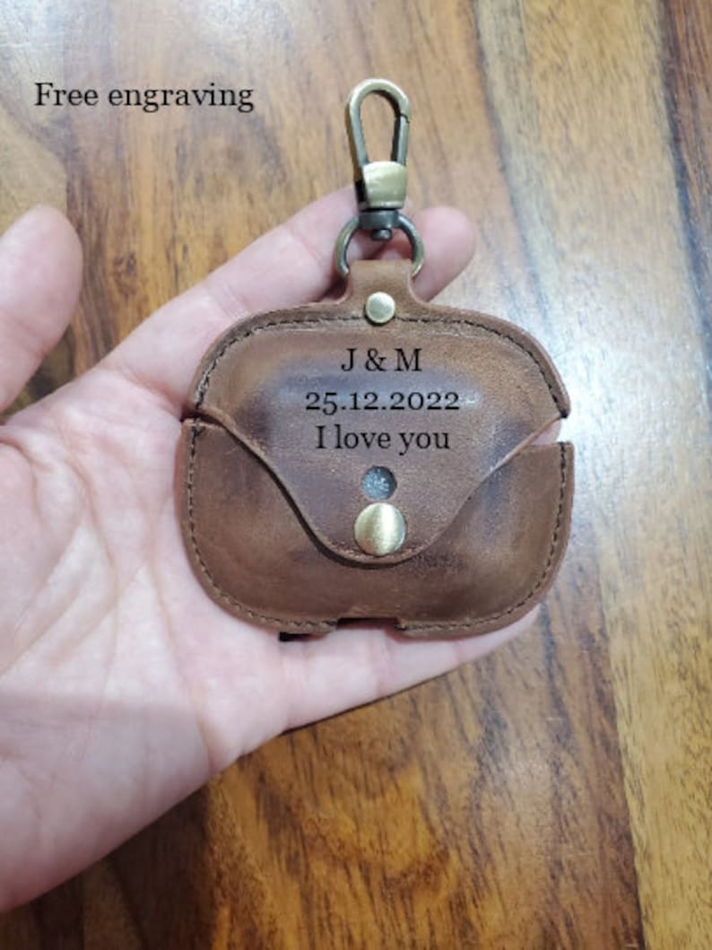 May include: A brown leather case with a gold metal clasp and a loop for attaching a keychain. The case has the initials "J & M" and the date "25.12.2022" engraved on it, along with the words "I love you".