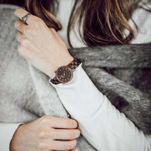 May include: A woman's wrist wearing a brown wooden watch with a rose gold face and black hands. The watch is on a white sweater and a gray fuzzy scarf.