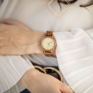 May include: A woman's wrist wearing a wooden watch with a light brown band and a white face. The watch has gold-coloured hands and a gold-coloured ring around the face.