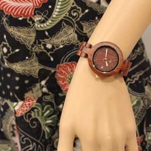 May include: A wooden wristwatch with a brown band and a round face. The watch face has a red wood grain pattern and gold-colored hands and hour markers.