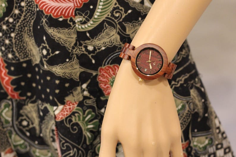 May include: A wooden wristwatch with a brown band and a round face. The watch face has a red wood grain pattern and gold-colored hands and hour markers.