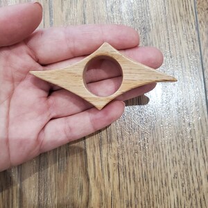 May include: A wooden bookmark shaped like a diamond with a round hole in the center. The bookmark is light brown in color.