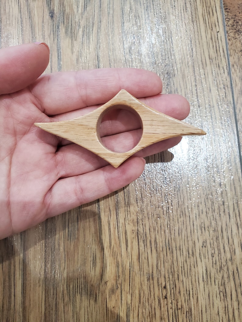 May include: A wooden bookmark shaped like a diamond with a round hole in the center. The bookmark is light brown in color.