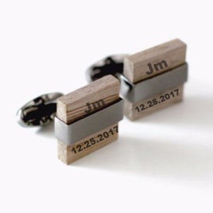 Op de afbeelding: Houten manchetknopen met een rechthoekig ontwerp. Elke manchetknop heeft de initialen "Jm" en de datum "12.25.2017" in zwart gegraveerd. De manchetknopen worden bij elkaar gehouden door een zilverkleurige metalen band.