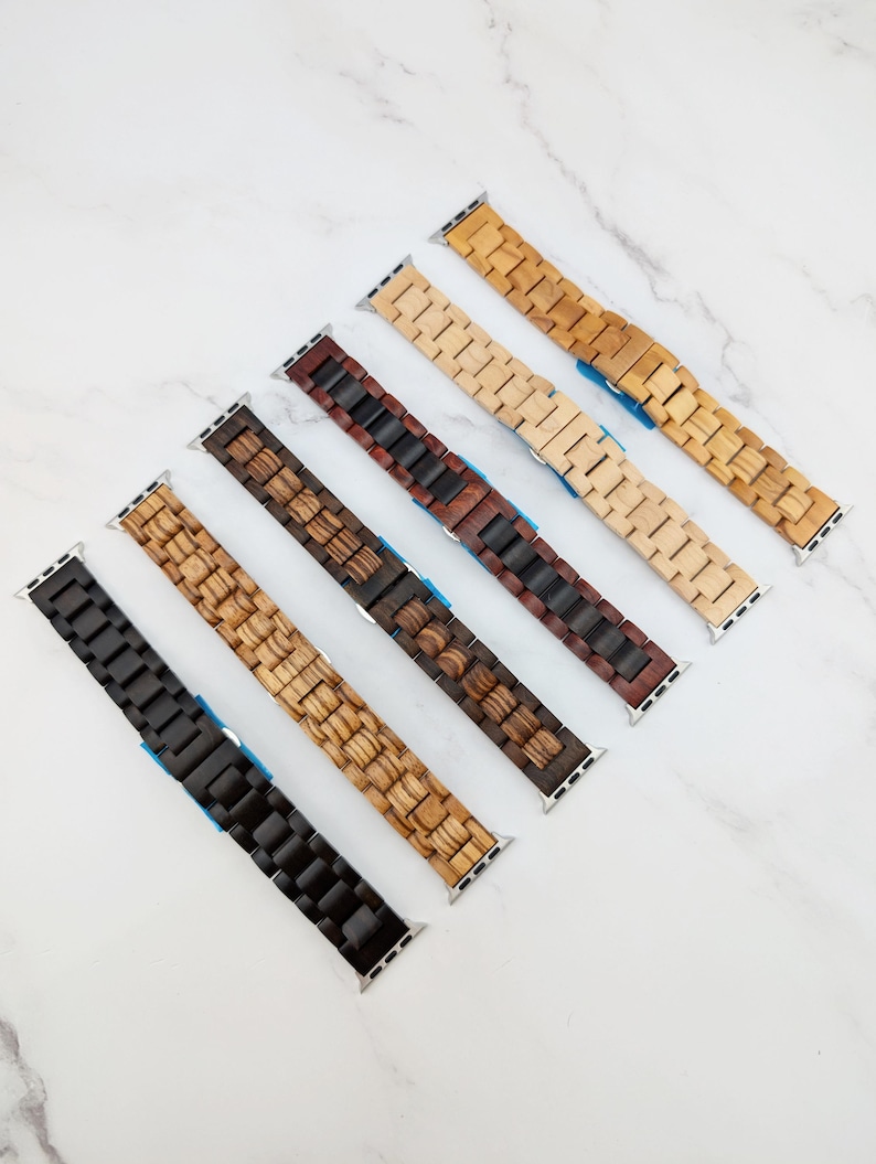 May include: Five different wood watch bands for Apple Watch. The bands are made of different types of wood, including light wood, dark wood, and a combination of both. The bands are all in a classic watch band style with a buckle closure.