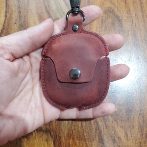May include: A burgundy leather case with a metal clasp and a metal loop for attaching a keychain. The case is designed to hold a pair of wireless earbuds.