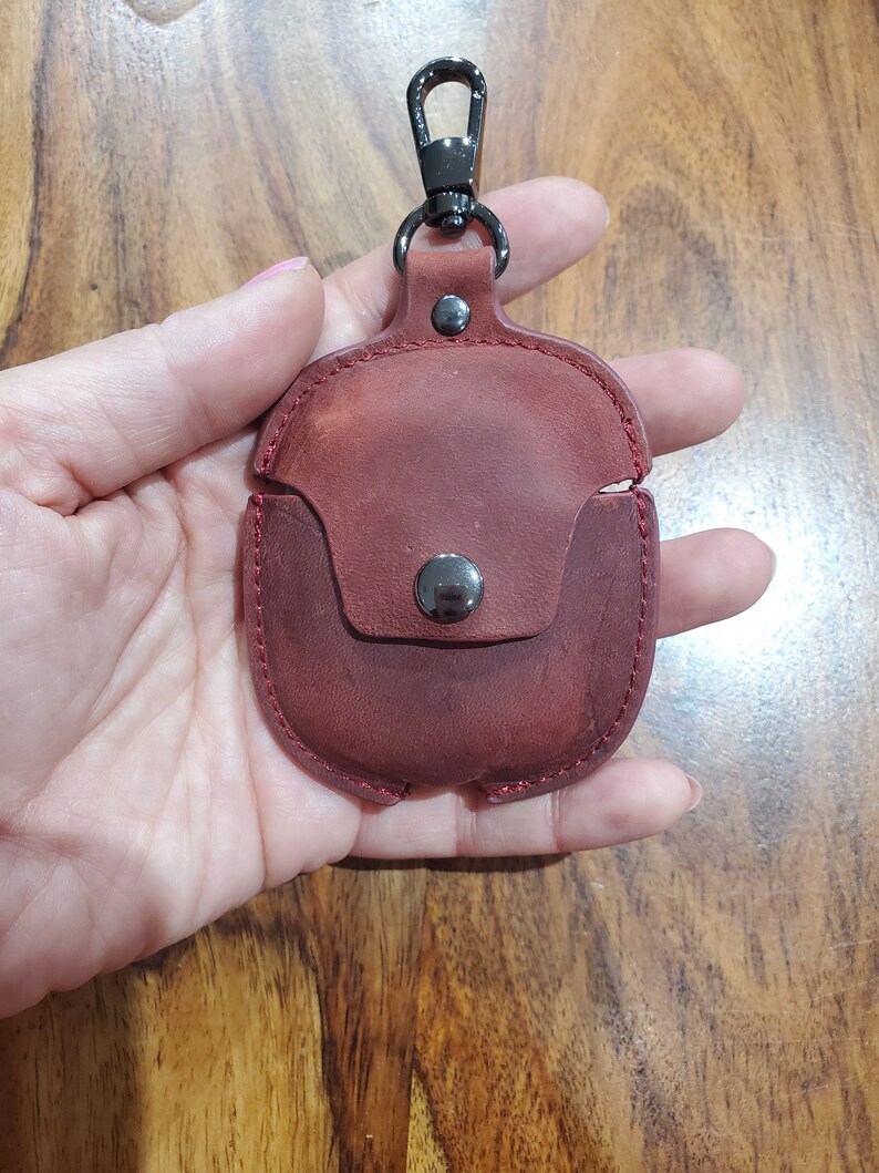 May include: A burgundy leather case with a metal clasp and a metal loop for attaching a keychain. The case is designed to hold a pair of wireless earbuds.