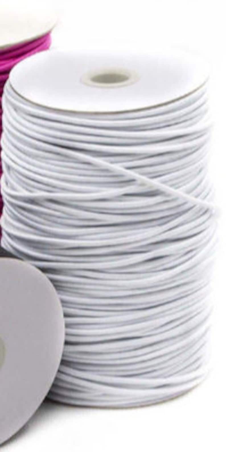 May include: A spool of white elastic cord. The cord is tightly wound around a white cardboard core. A partial view of a pink spool is visible in the background. The elastic cord is used for sewing and crafting projects.