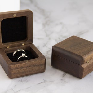 Könnte beinhalten: Zwei Holz-Ringboxen, eine offen und eine geschlossen, liegen auf einer Marmoroberfläche. Die offene Box zeigt zwei Ringe, die in schwarzem Samt liegen. Die Boxen sind aus dunklem Holz mit quadratischer Form und Messingscharnieren.