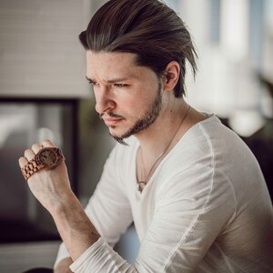 May include: A man wearing a white long-sleeved shirt and black pants is looking down at a wooden wristwatch with a brown band.