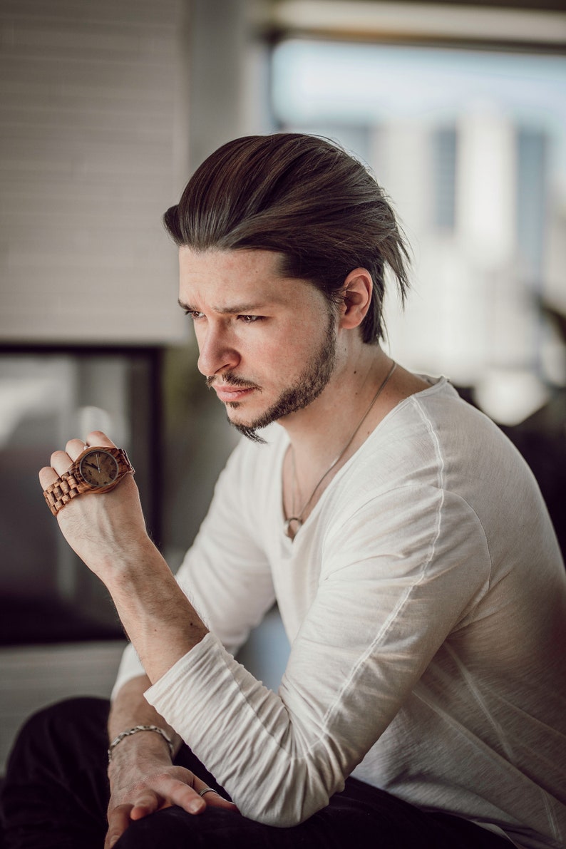May include: A man wearing a white long-sleeved shirt and black pants is looking down at a wooden wristwatch with a brown band.